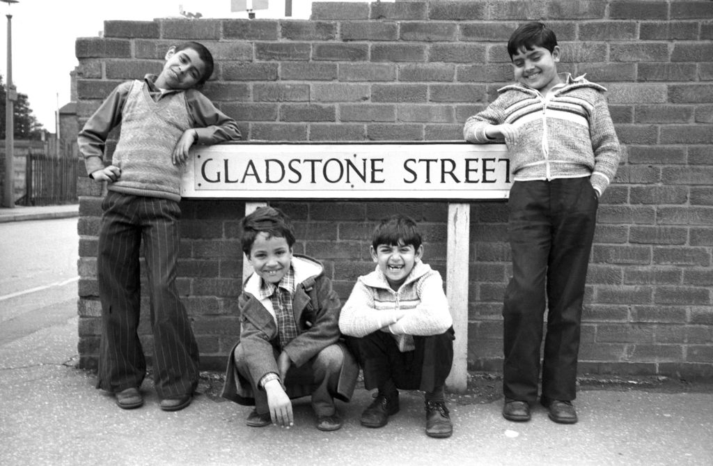 A black & white photograph of four South Asian boys stood around a Gladstone Street sign. Taken in 1970s.