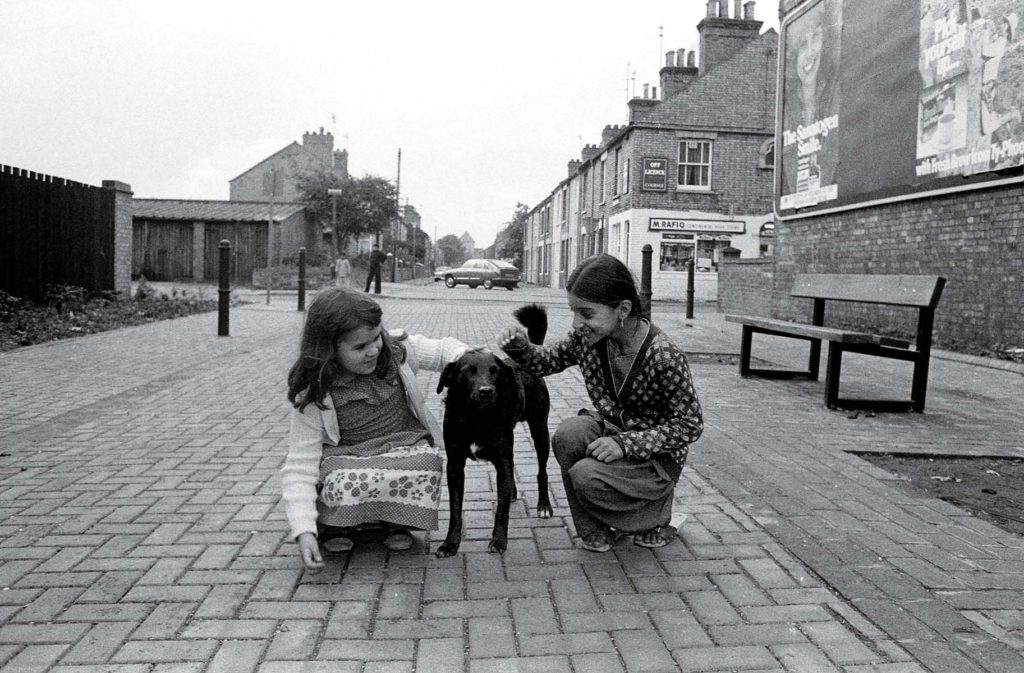 A black and white streetview image of 2 little girls sitting down to pet a dog